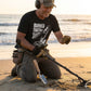 Man on a beach using a metal detector with headphones, wearing a 'Buried Truth Seeker' t-shirt.