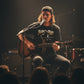 Person playing guitar on stage with a microphone, wearing a 'Born to Play' shirt.